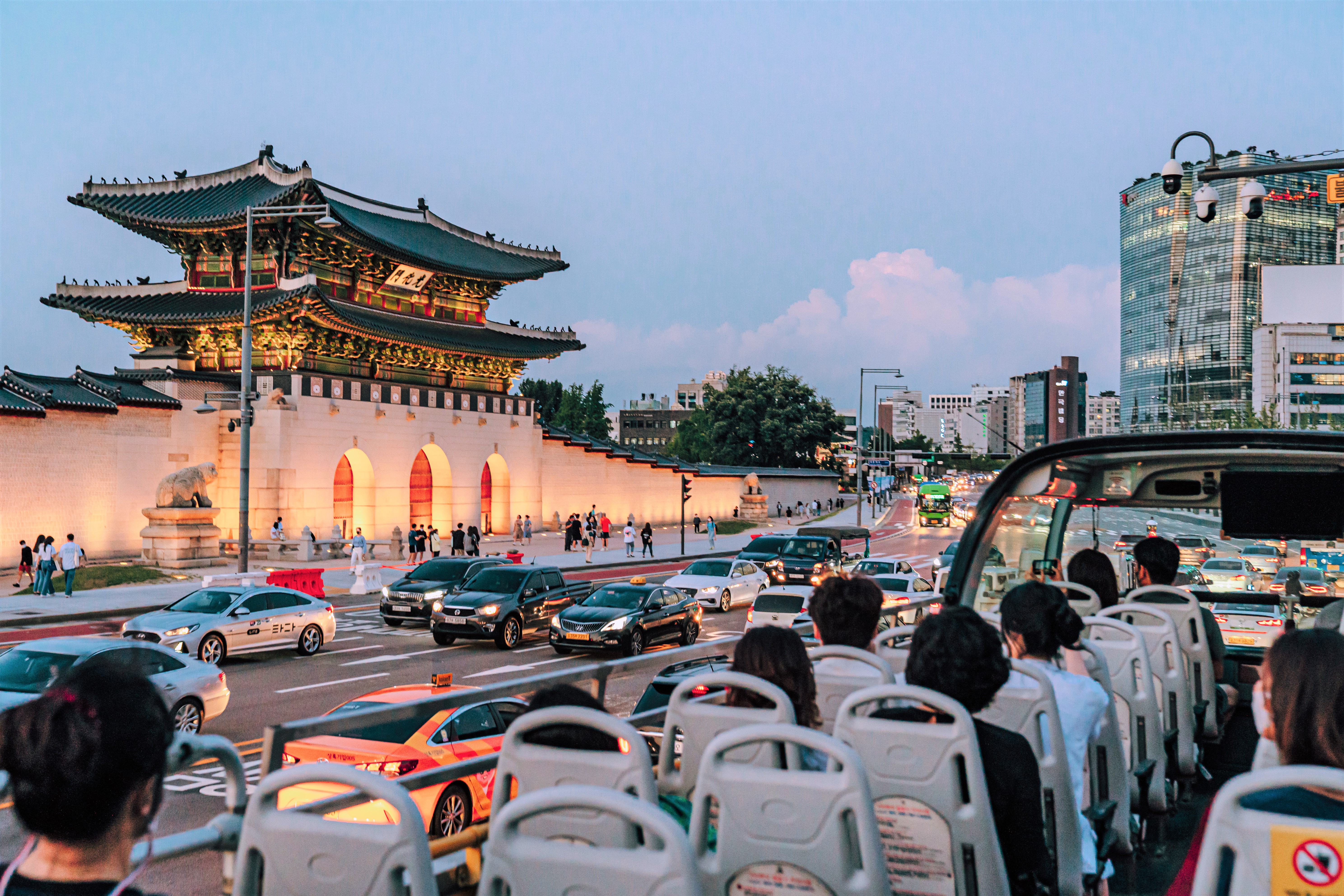 Seoul City Tour Bus driving on a road in downtown Seoul, people are riding the Seoul City Tour Bus, cars are on the road, and Gwanghwamun Gate is seen with its lights on