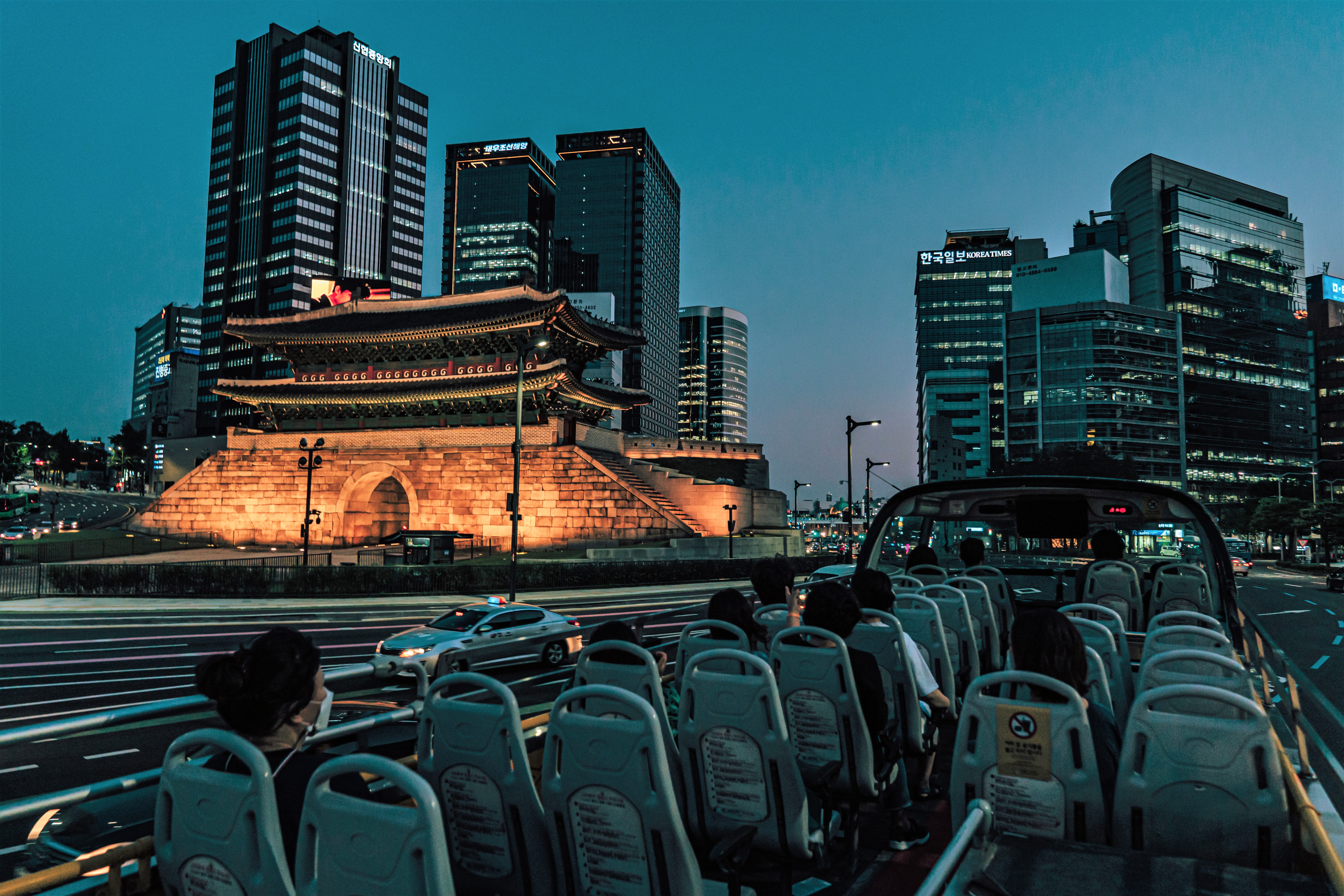 Seoul City Tour Bus driving on the road in downtown Seoul, Sungnyemun Gate and downtown Seoul are in the background, and there are people on the Seoul City Tour Bus.