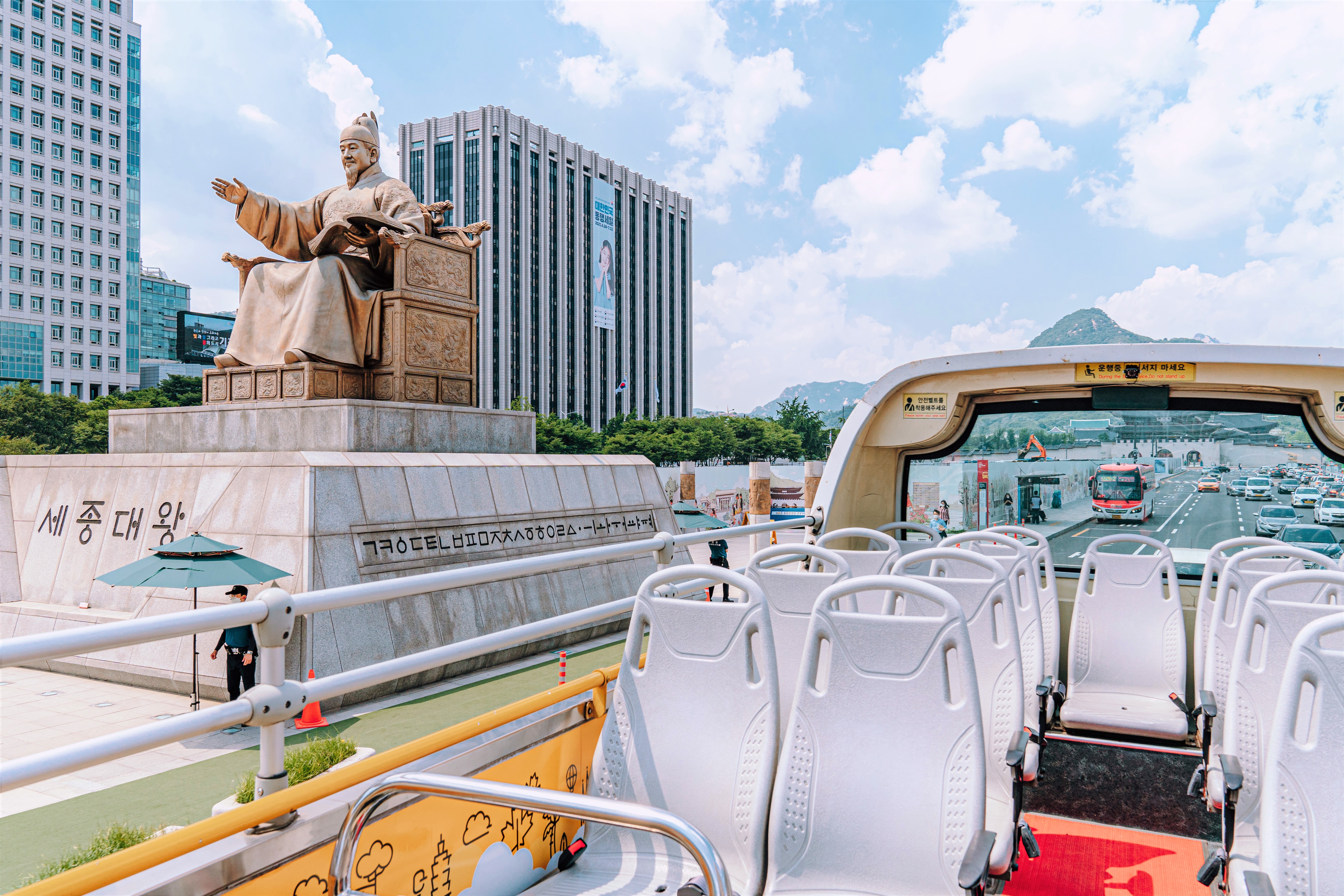 The statue of King Sejong seen from the Seoul City Tour Bus. There are beautiful clouds in the sky and cars driving on the road.