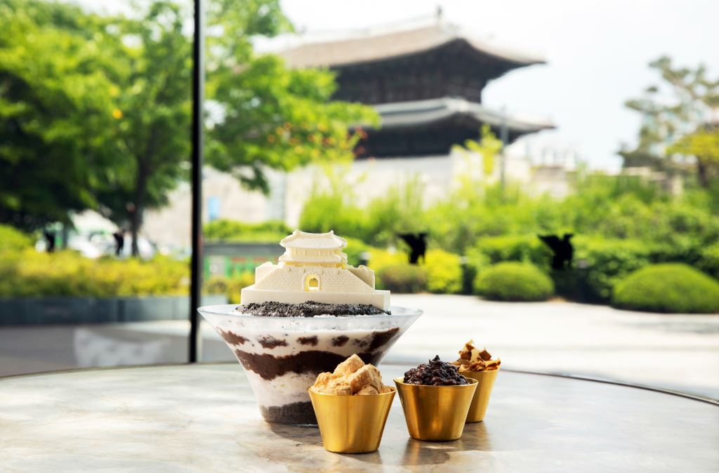 Gambaran Bingsu yang dihiasi dengan coklat yang berbentuk Heunginjimun beserta 3 mangkuk kecil yang berisi isi tambahan. Terlihat juga gerbang Heunginjimun di latar.