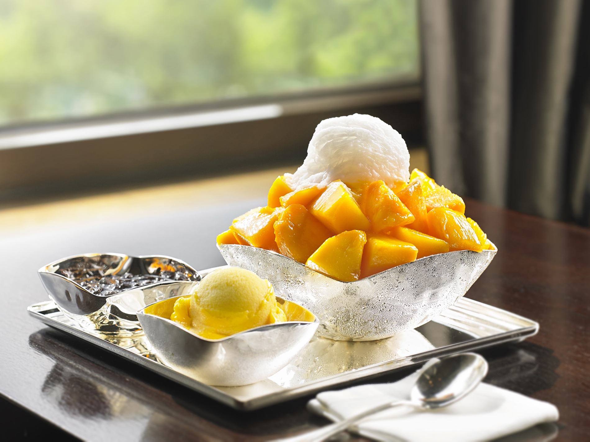 There is shaved ice topped with mango, and there is a small bowl and spoon with mango ice cream right next to it.