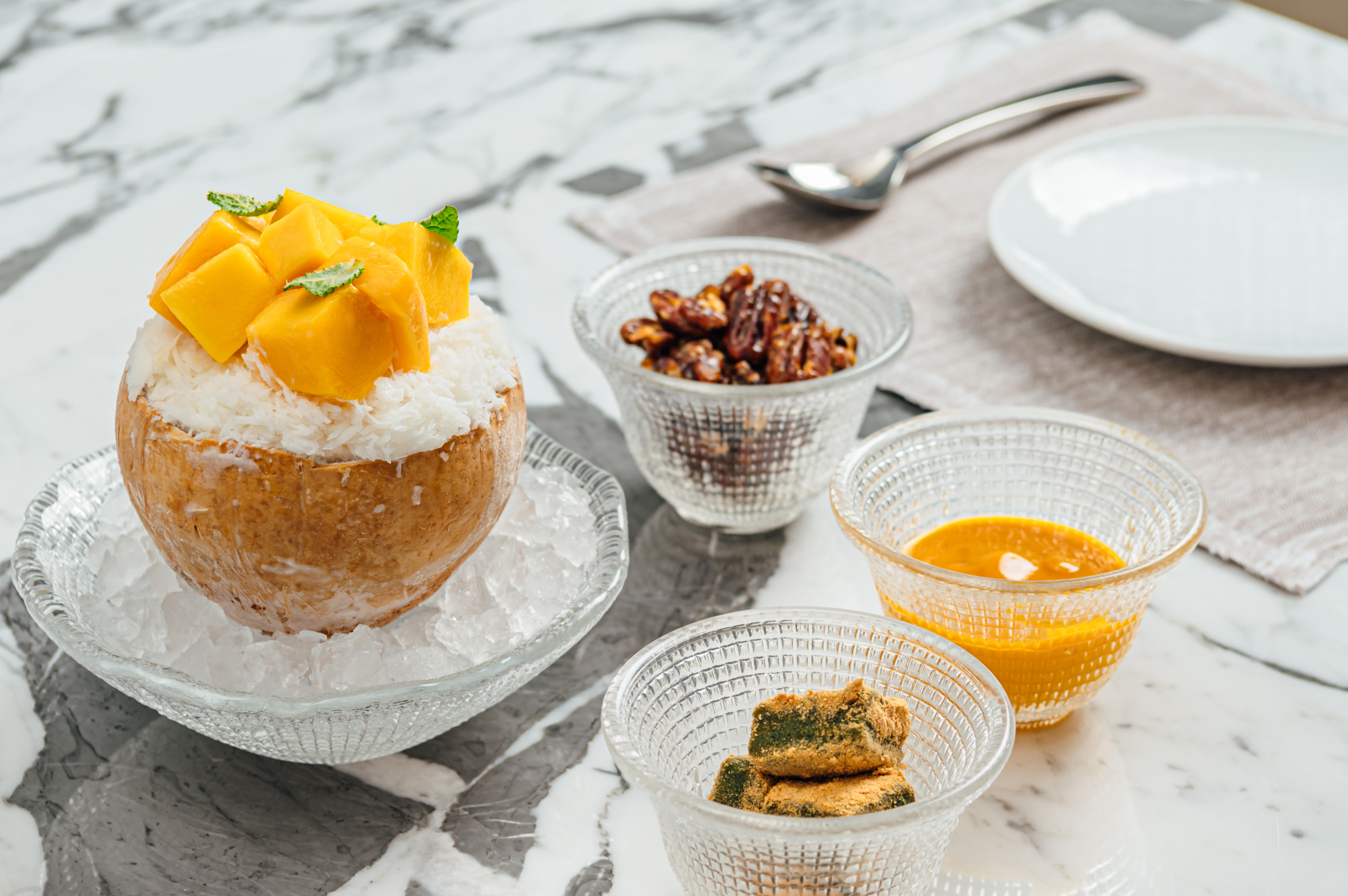 Coconut mango shaved ice is sitting in a coconut shell, and three small bowls with toppings are placed around it.