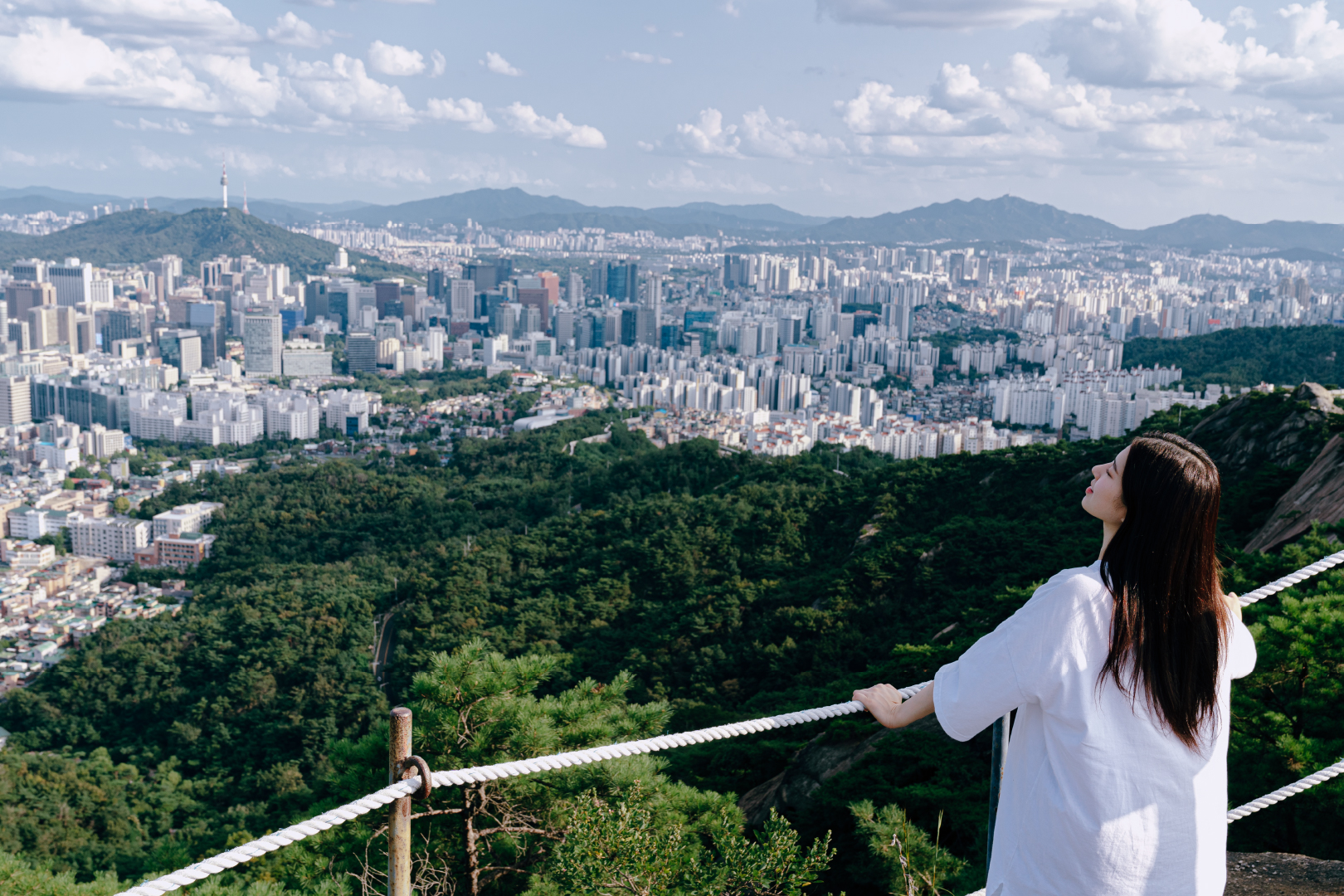 흰색 옷을 입은 여자가 산에서 서울 전망을 보고 있다.