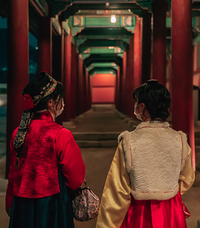 Dua wanita yang memakai hanbok sedang berjalan di koridor berlatarkan tiang yang terbaris rapi.